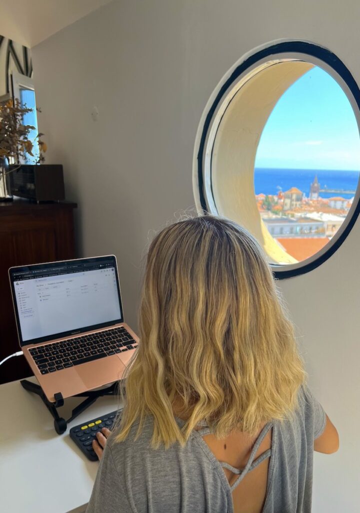 Blonde woman types on laptop at a desk next to a circular window with a view of Funchal in Madeira.