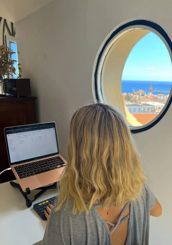 Blonde woman types on laptop at a desk next to a circular window with a view of Funchal in Madeira.