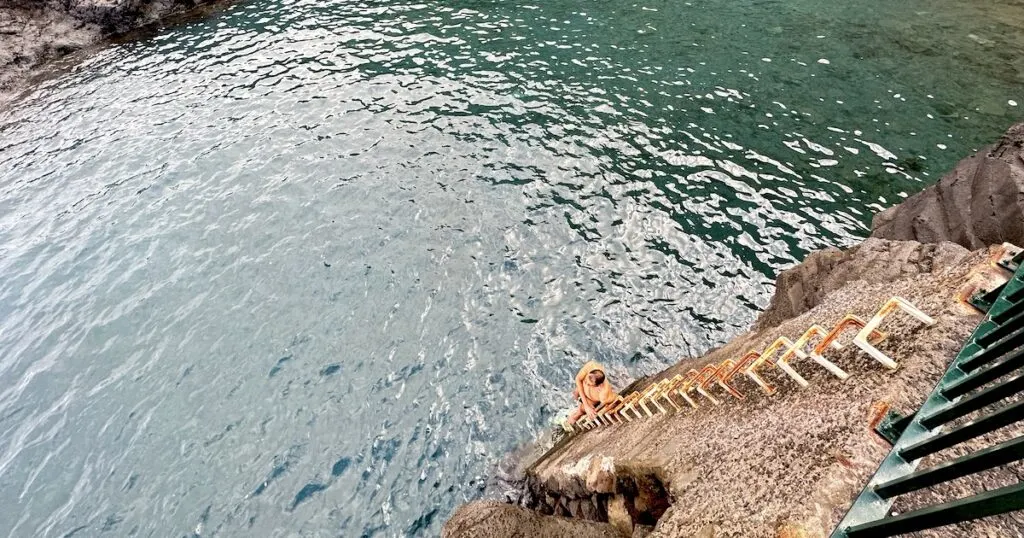 A man climbs from the pool onto a rusty ladder stapled into the wall.