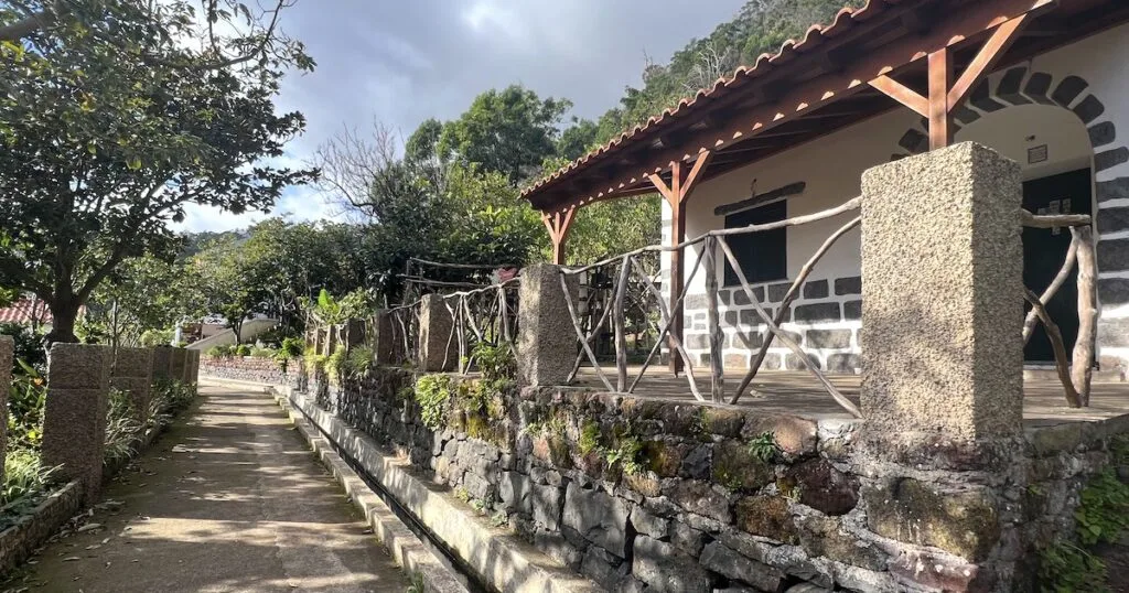 The beginning of the Levada dos Maroços trail in Machico, passing by a stone house with beams.