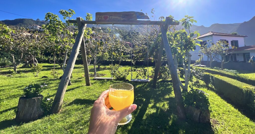 A hand holds a glass of orange poncha in front of a swing set at Granny's Farm in Madeira.