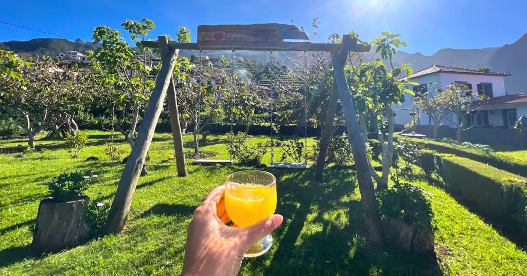 A hand holds a glass of orange poncha in front of a swing set at Granny's Farm in Madeira.