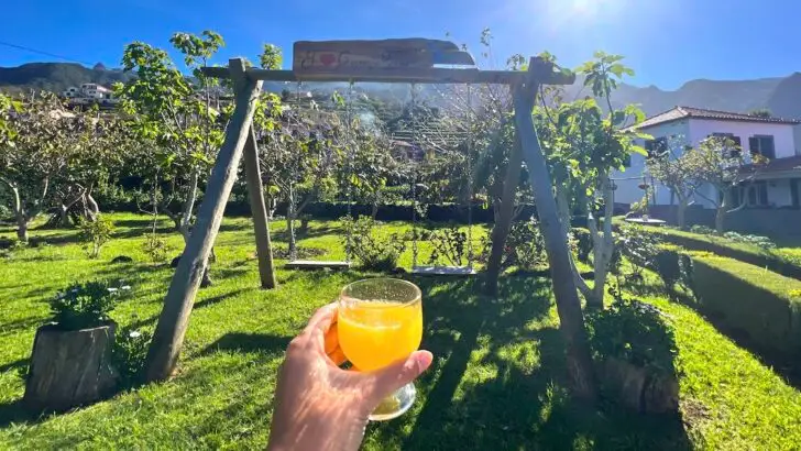 A hand holds a glass of orange poncha in front of a swing set at Granny's Farm in Madeira.