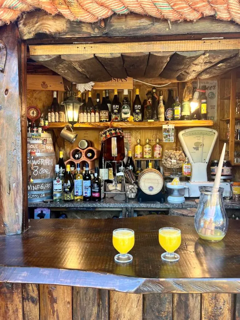 Two glasses of poncha and a large jar on a wooden bar with rows of bottles.
