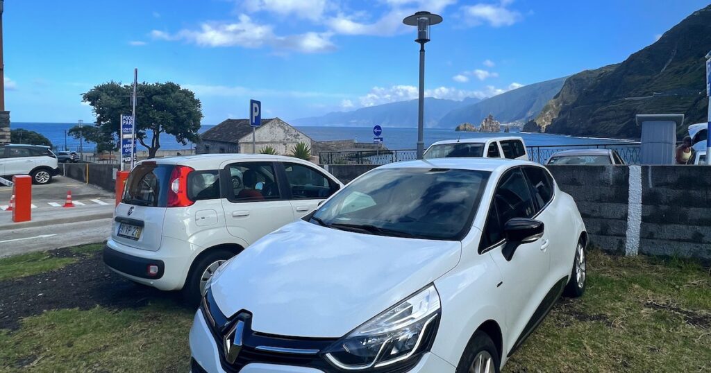 Two small, white cars are parked in a car park overlooking the sea in Porto Moniz.