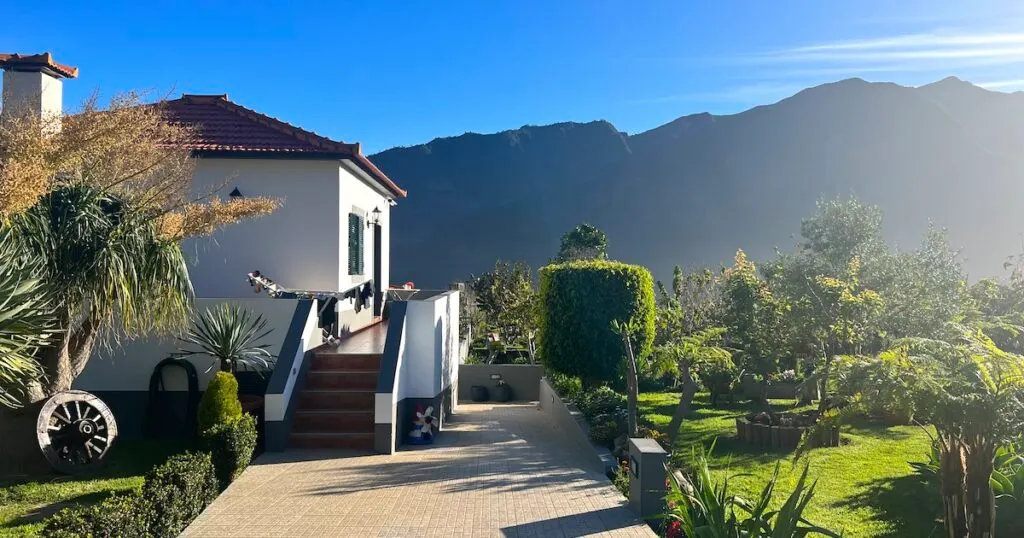 A white cottage next to landscaped gardens, with mountains in the background.