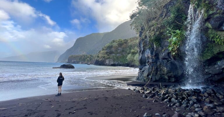 A woman wearing a hoodie and shorts gazes at a rainbow over the ocean while a waterfall pours from the cliff at Seixal Beach in Madeira.