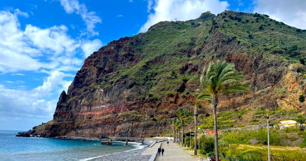 Tall green cliffs and palm trees tower over a concrete promenade in Madalena do Mar, Madeira, next to the Atlantic Ocean.