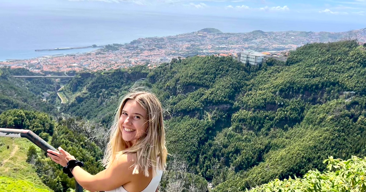 Woman smiling at a viewpoint above Funchal on Levada dos Tornos, with lush green valleys and the Atlantic coastline in the background.
