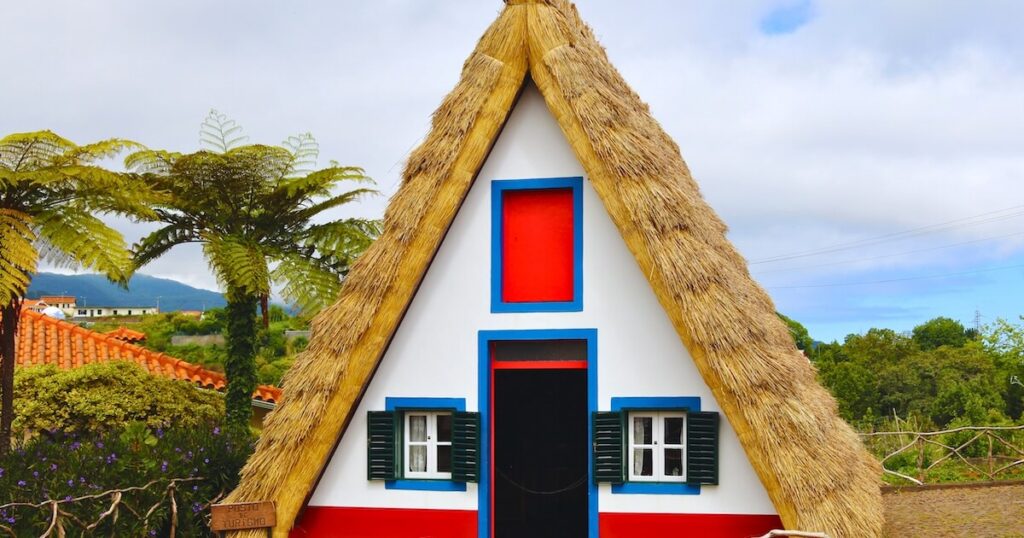 Traditional Madeira Santana house with a steep thatched roof, white walls and bright red and blue trim surrounded by tropical greenery.