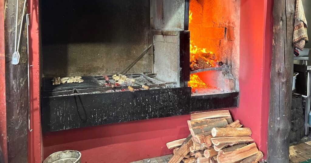 Wood-fired barbecue grill with skewers cooking beside an open flame and stacked logs ready for the fire.