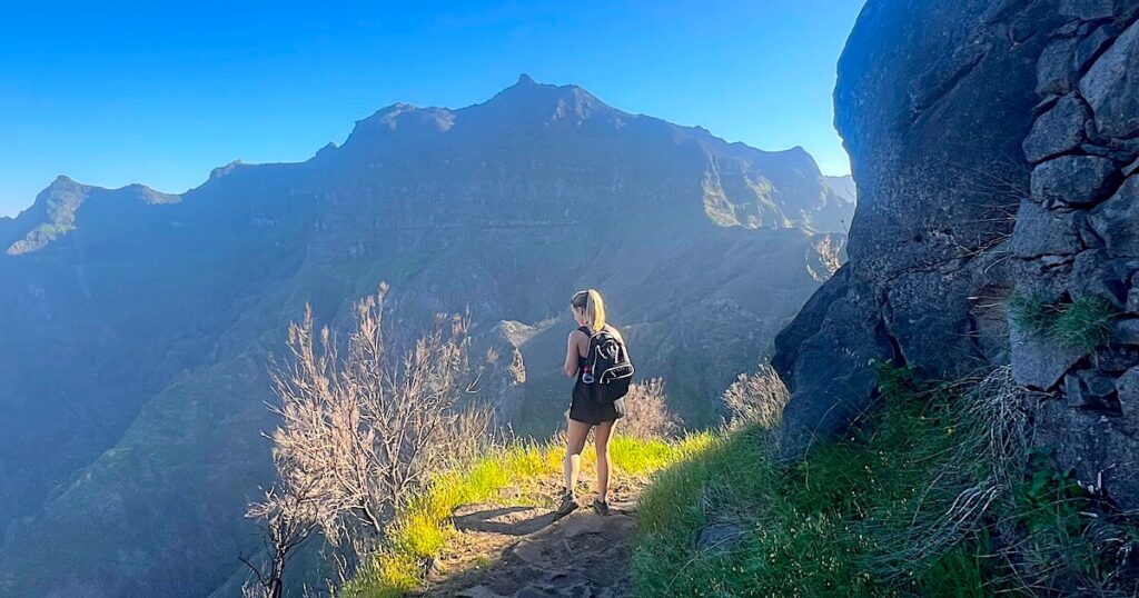 Hiker standing on a narrow cliffside path (the PR12 to Pico Grande) with panoramic views of Madeira’s mountains.