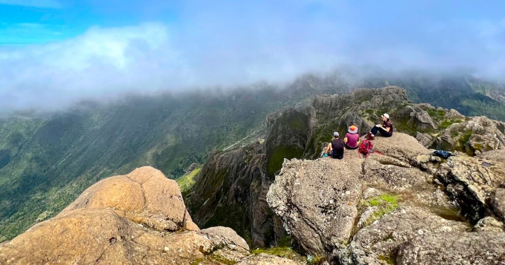 Hikers resting on a rocky summit ledge above deep valleys and drifting cloud.