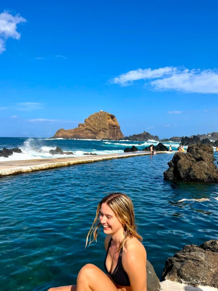 Woman sitting beside a natural volcanic swimming pool on Madeira’s coast, with waves breaking against black volcanic rocks.