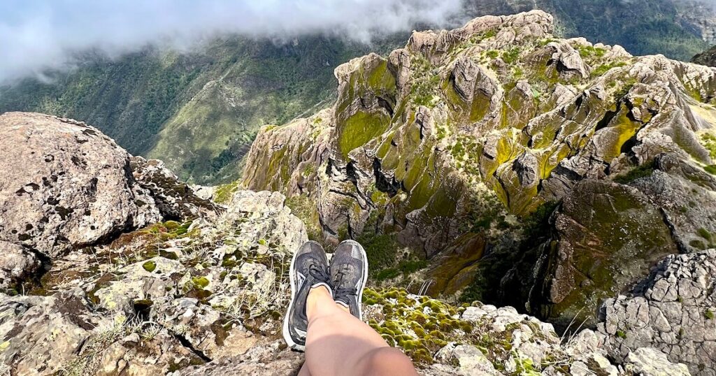 Hiker’s feet dangling over a rocky cliff edge overlooking the Pico Grande ridgeline.