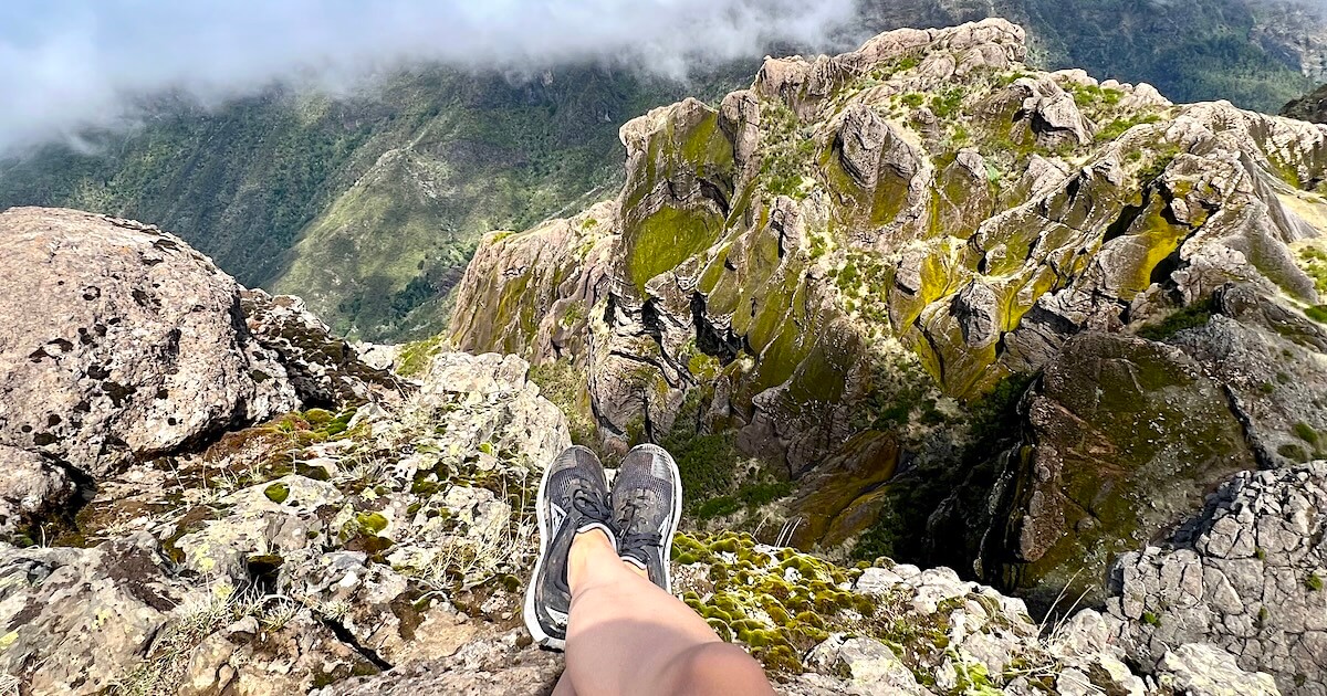 Hiker’s feet dangling over a rocky cliff edge overlooking the Pico Grande ridgeline.