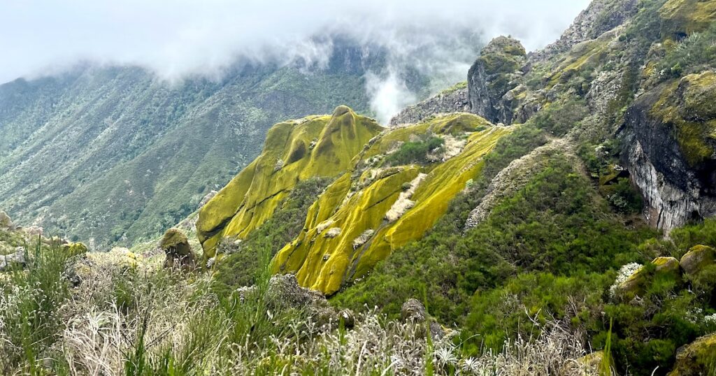 Vivid green moss-covered rock formations emerge from a rugged mountainside as low clouds drift across the steep valley beyond.