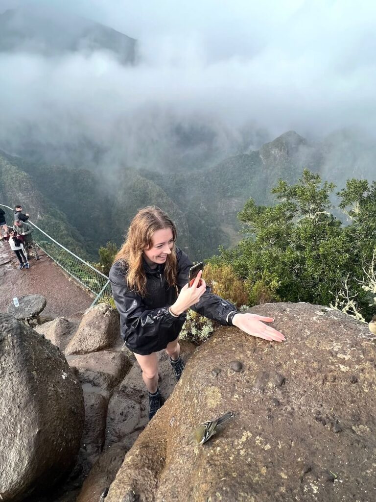 Woman photographing a small bird on a rock with cloud-covered Madeira mountains behind her.