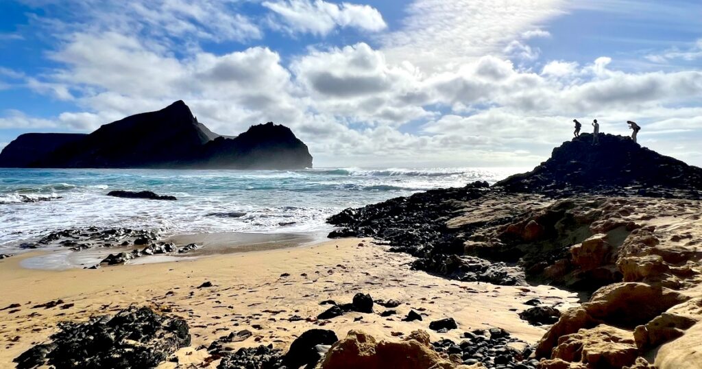 Three shadowy figures stand on a rock at the sandy beach at Ponta da Calheta in Porto Santo.