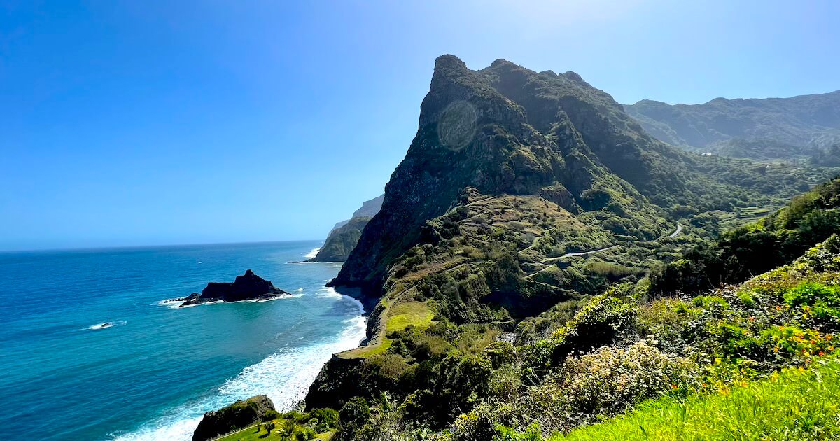 Green coastal cliffs and a winding hillside road above the blue Atlantic, seen from São Cristóvão viewpoint in Madeira.