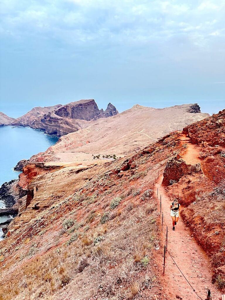 Hiker following a narrow red-earth path beside a safety fence above the sea at Ponta de São Lourenço.