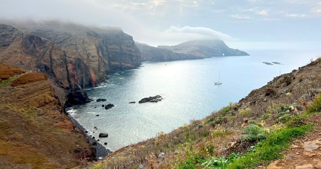 Mist covering the tops of rugged cliffs above a calm bay at Ponta de São Lourenço.