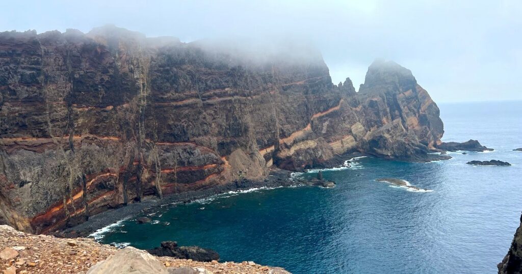 Layered orange and dark volcanic cliffs rising above blue water, with mist obscuring their tops.