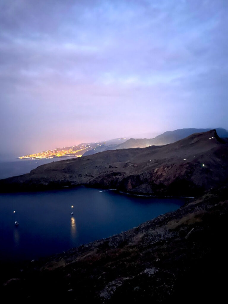 Sheltered bay and dark coastal ridges along Madeira’s PR8 trail before sunrise, with distant lights beneath a purple sky.