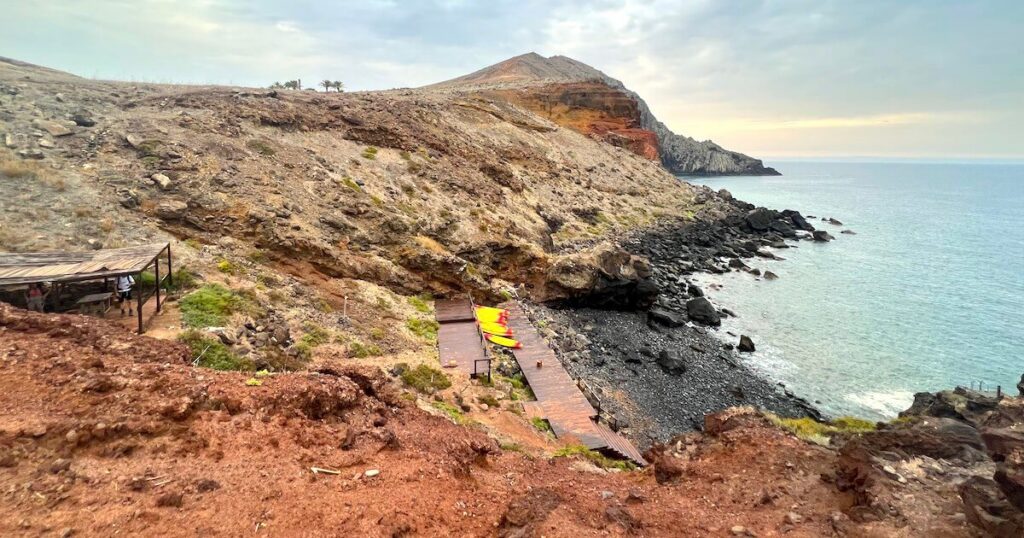 Small pebble beach below dry coastal slopes at Praia do Sardinha, with a yellow kayak near the shore.