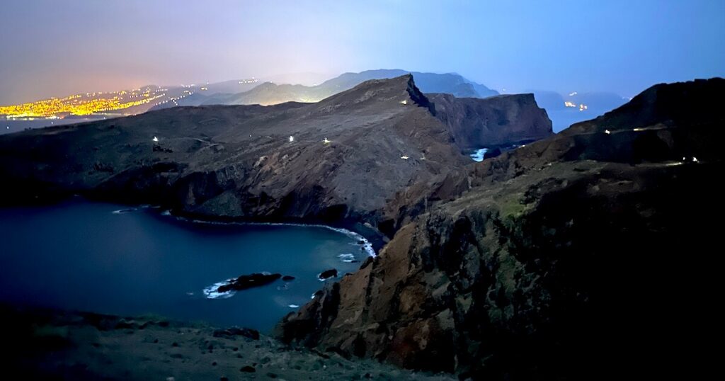 Rocky coastline and sheltered bay along the PR8 trail in low light, with distant lights on the horizon.