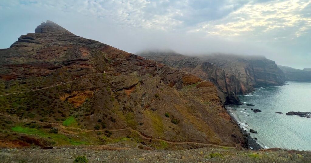 Narrow walking trail crossing a rugged coastal ridge above the sea, with mist covering distant cliffs.